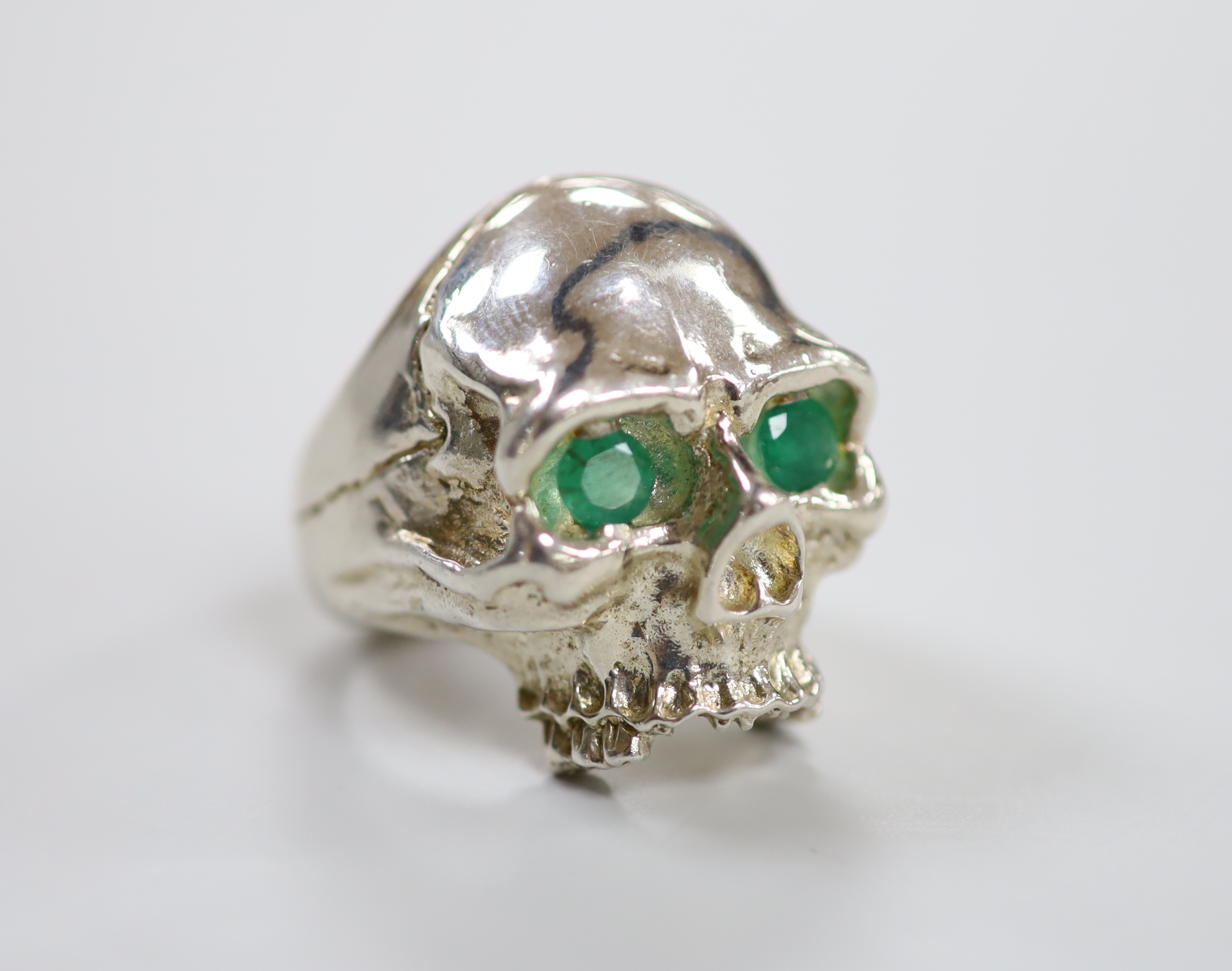 A white metal (tests a silver) and two stone emerald set skull ring, size T/U, gross weight 35,2 grams.